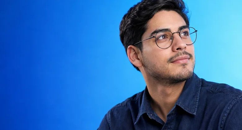 Hombre joven con lentes redondos de metal negro mira a lo lejos sobre un fondo azul.